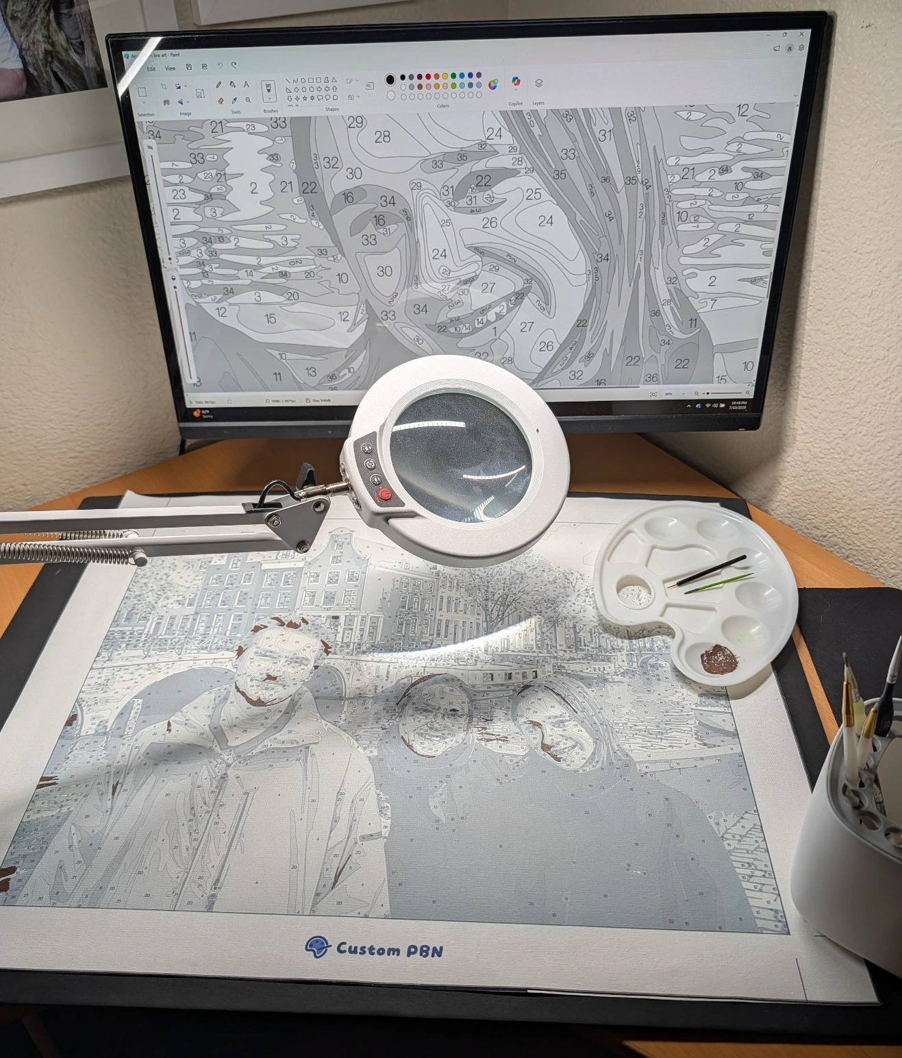 A CustomPBN paint by number canvas under a magnifying lamp, with a zoomed-in HD digital reference file displayed on a computer monitor in the background.