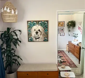 A framed custom pet portrait of a white fluffy dog hanging on a white wall in a stylishly decorated room with plants and modern furniture.