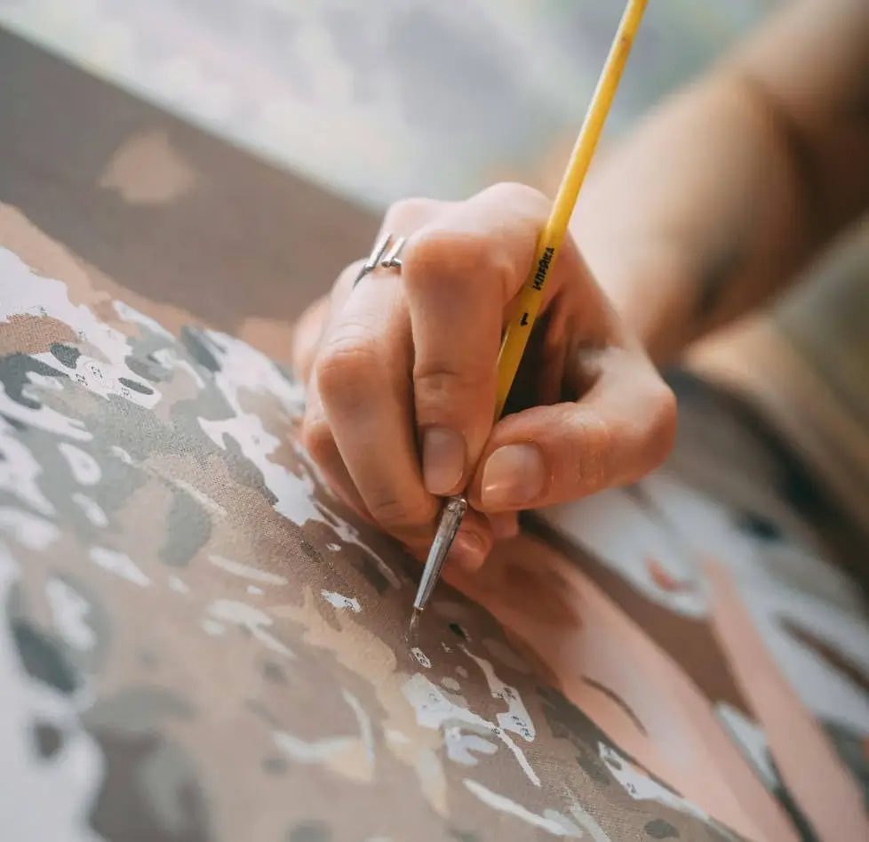 A close-up of a hand carefully painting a custom paint by numbers canvas with a yellow brush, highlighting the relaxing process.